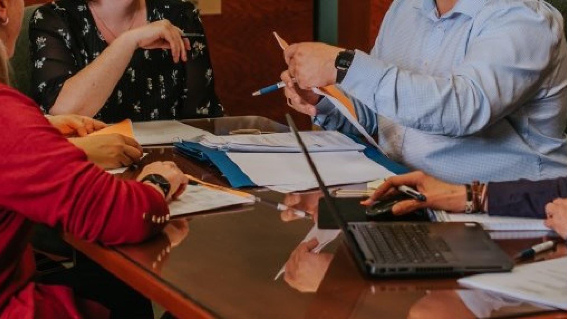 Group of people having a meeting at a wooden table with laptops and papers.