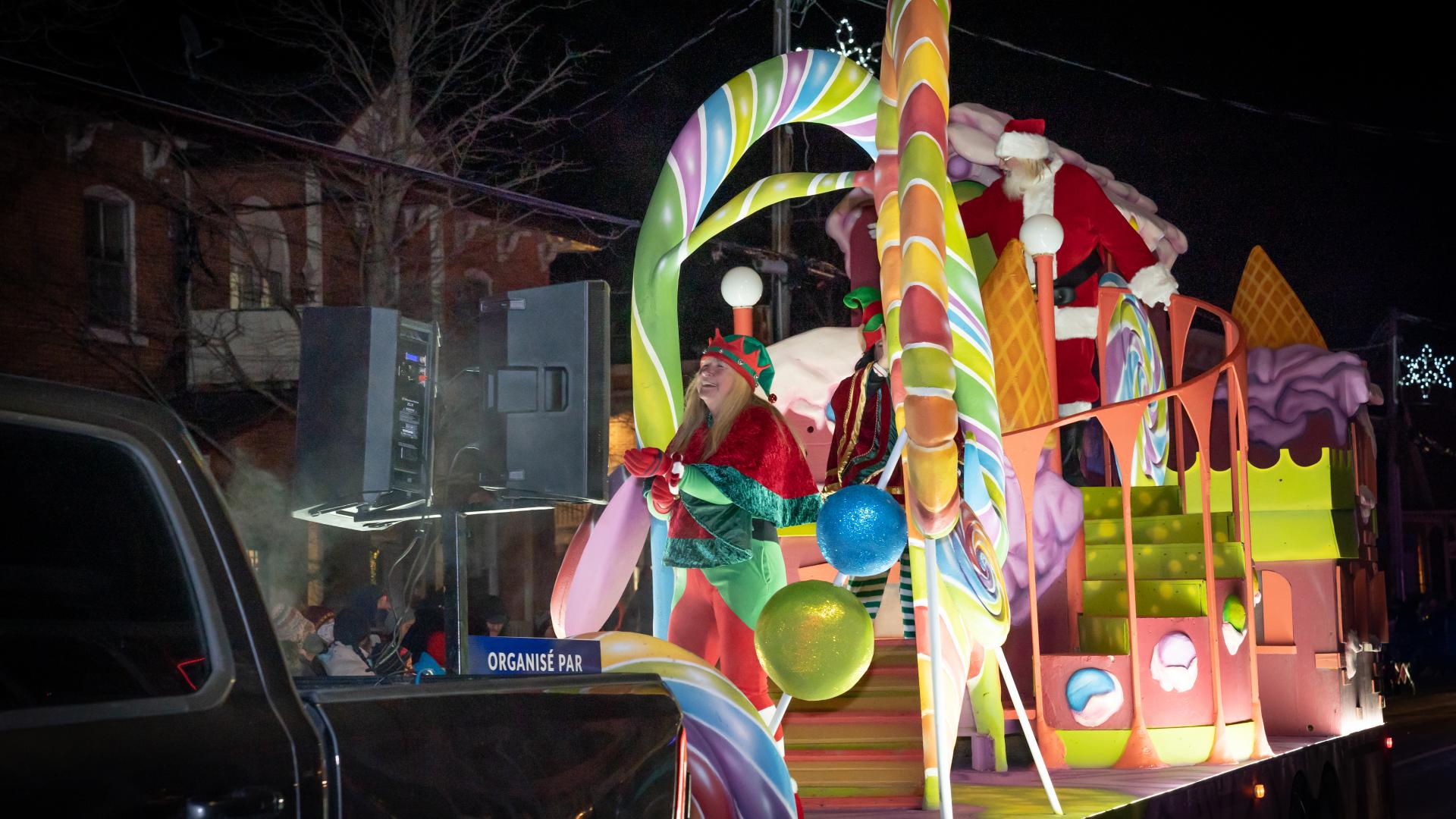 A colourful candy-themed Christmas parade float featuring Santa Claus, elves, and oversized candy decorations at night.