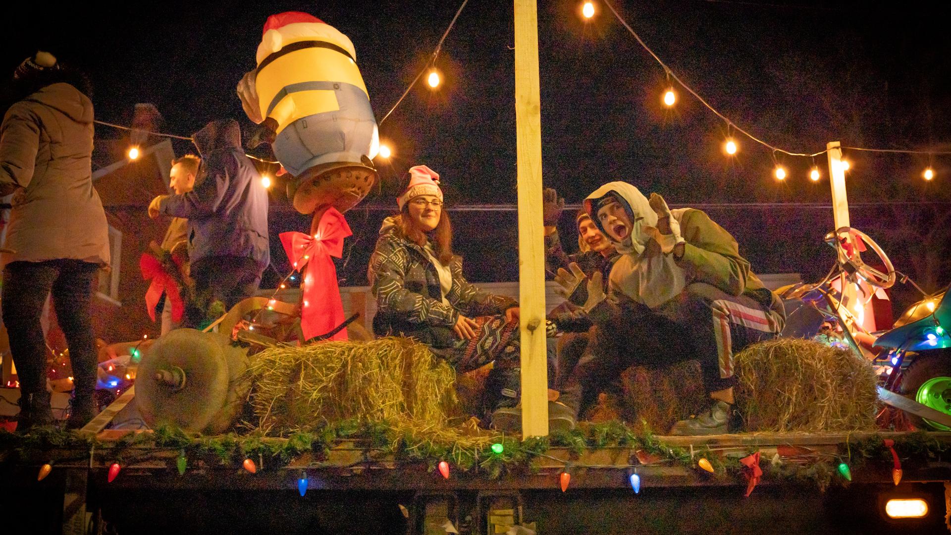 Community members riding on a festive float decorated with string lights, hay bales, and holiday decorations during the Christmas parade.