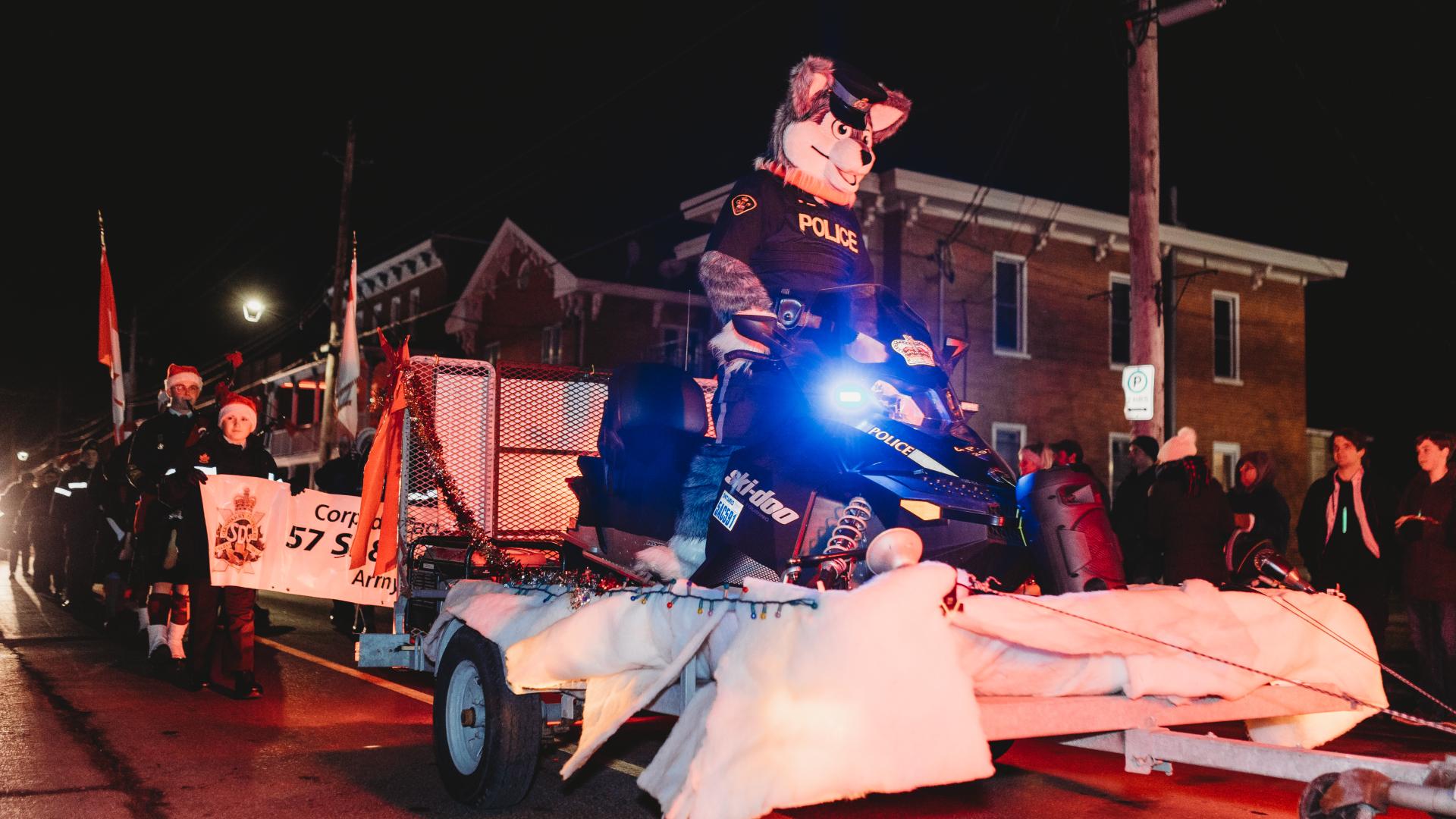 An OPP mascot riding a snowmobile in the Christmas parade with community members walking behind carrying a banner.