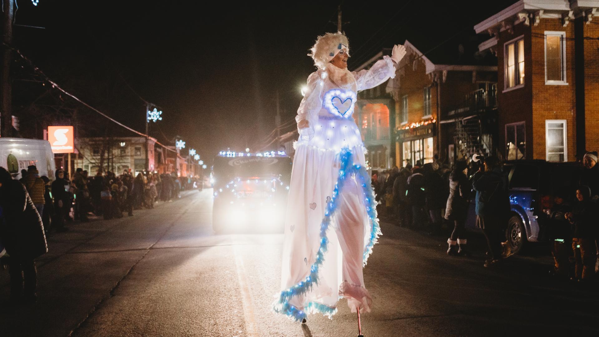 Performer dressed in a glowing winter costume waving to spectators during the Christmas parade at night.