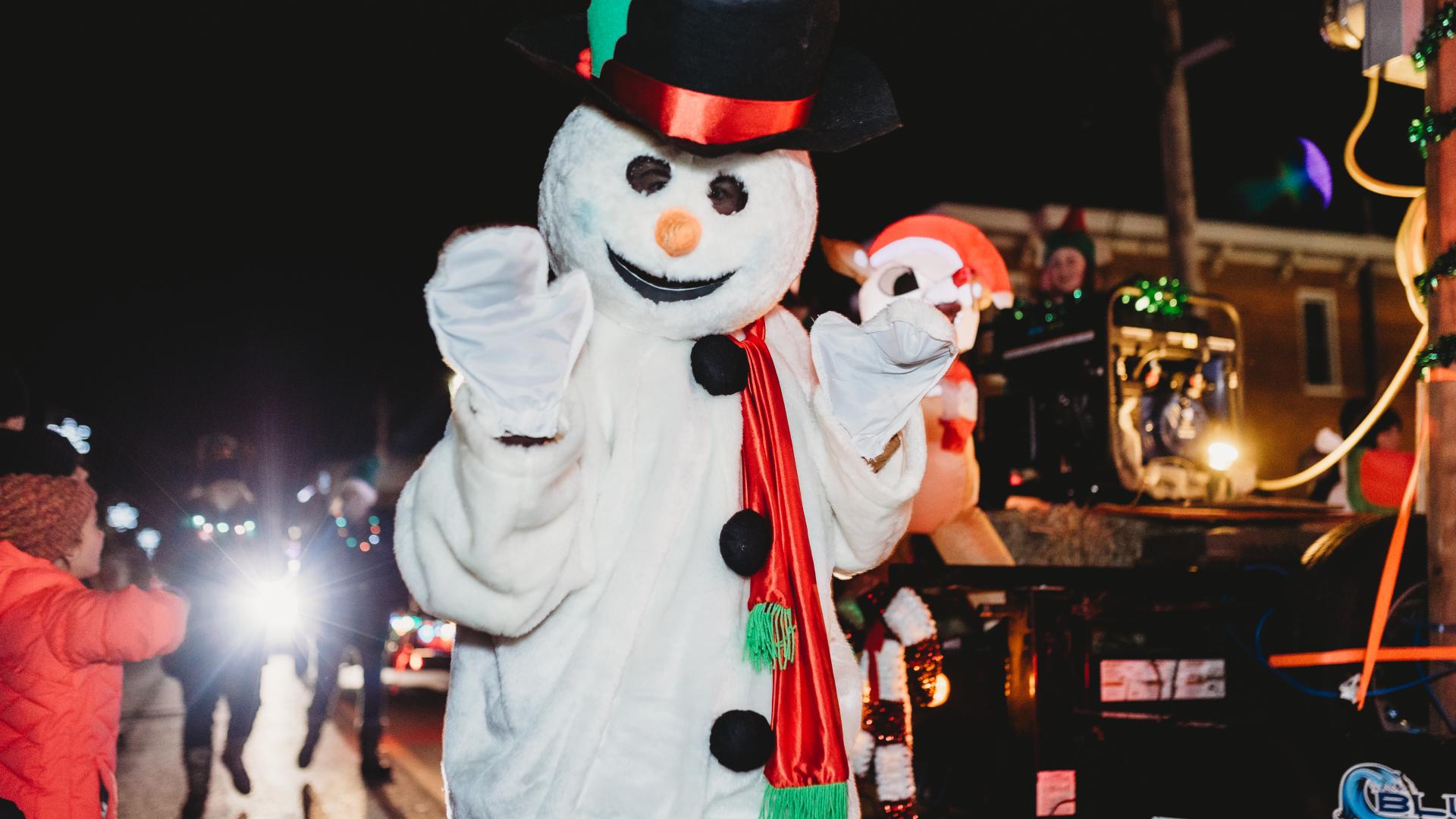 A snowman mascot waving to spectators during the nighttime Christmas parade celebrations.