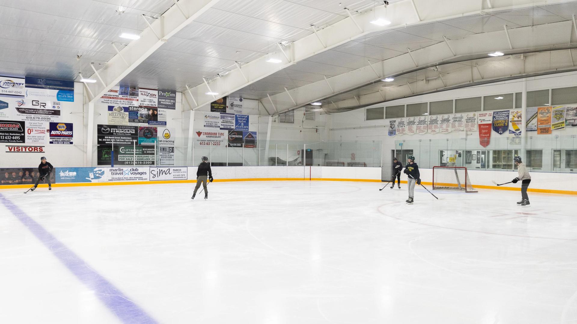 Wide view of the Vankleek Hill Arena ice surface with skaters on the ice and hockey boards surrounding the rink.