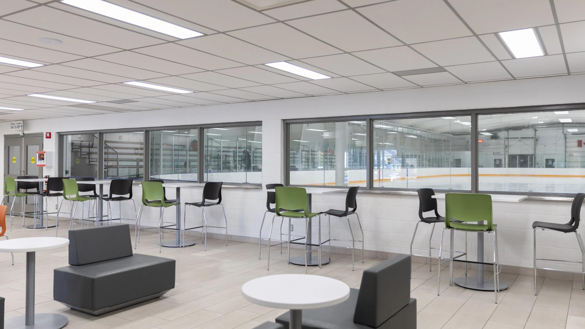 Bright arena lounge and viewing area with tables, chairs, and large windows overlooking the ice rink.