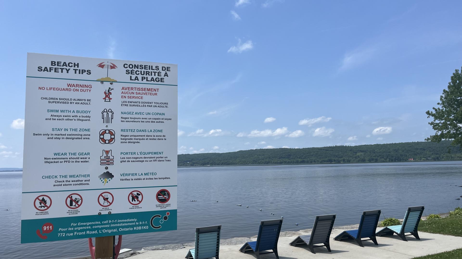 Beach safety rules sign beside the Ottawa River, with lounge chairs and the waterfront visible in the background.