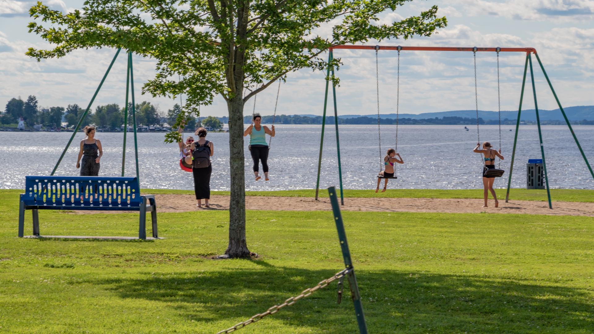 Children playing on swings in L'Orignal Park, with the Ottawa River and open green space visible in the background.
