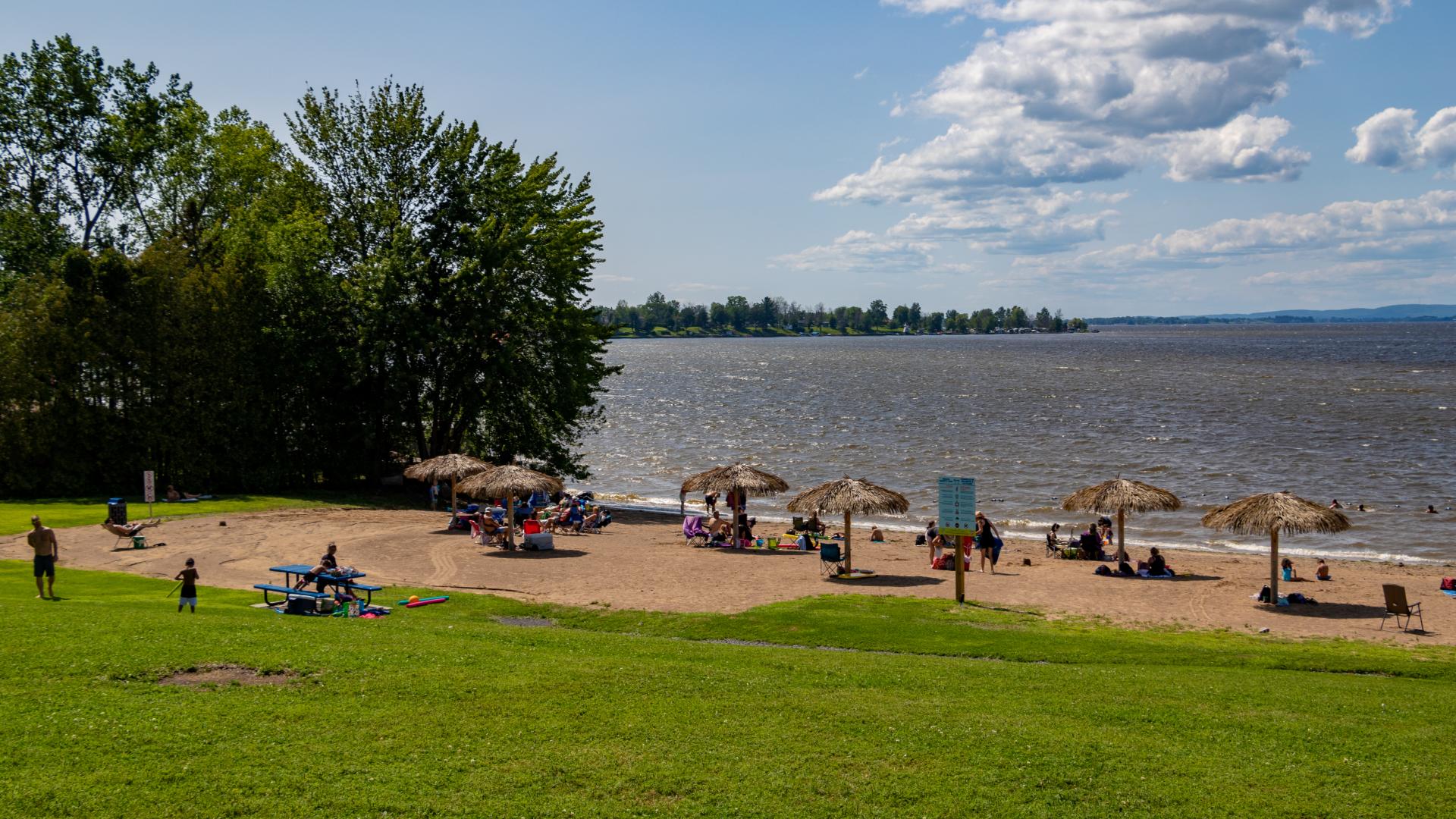 People enjoying a sandy beach along the Ottawa River, with lounge chairs, picnic areas, and waterfront views on a sunny day.