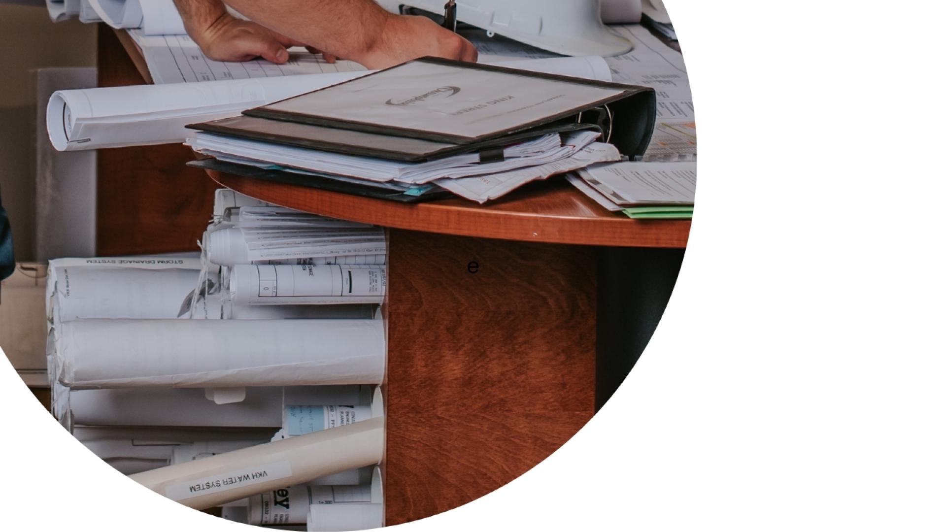 Office desk with rolled construction drawings, binders, documents, and a hand organizing paperwork