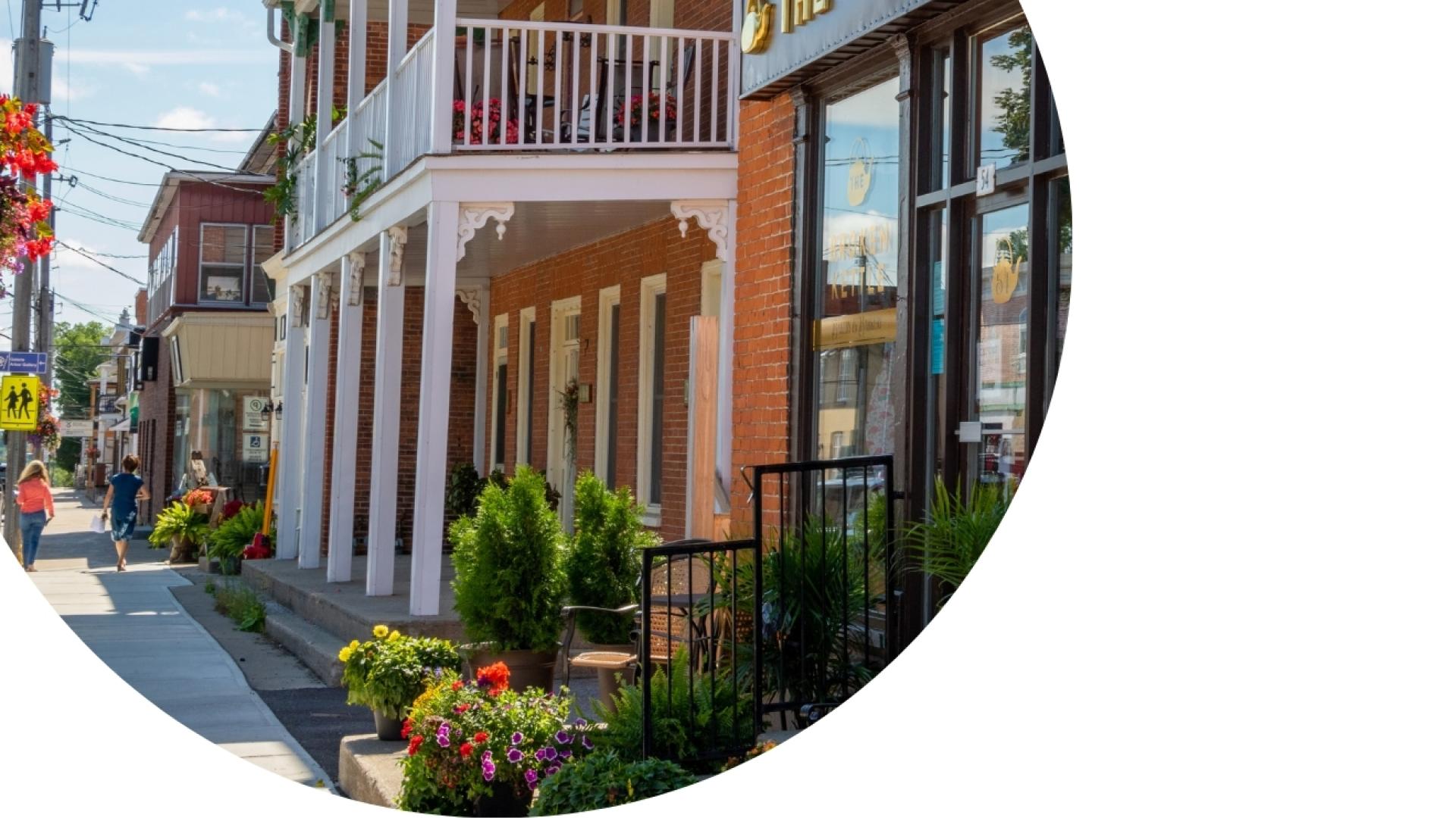 Streetscape in downtown Vankleek Hill featuring historic brick storefronts, colourful flower planters, sidewalk patios, and pedestrian-friendly streetscape elements along Main Street.