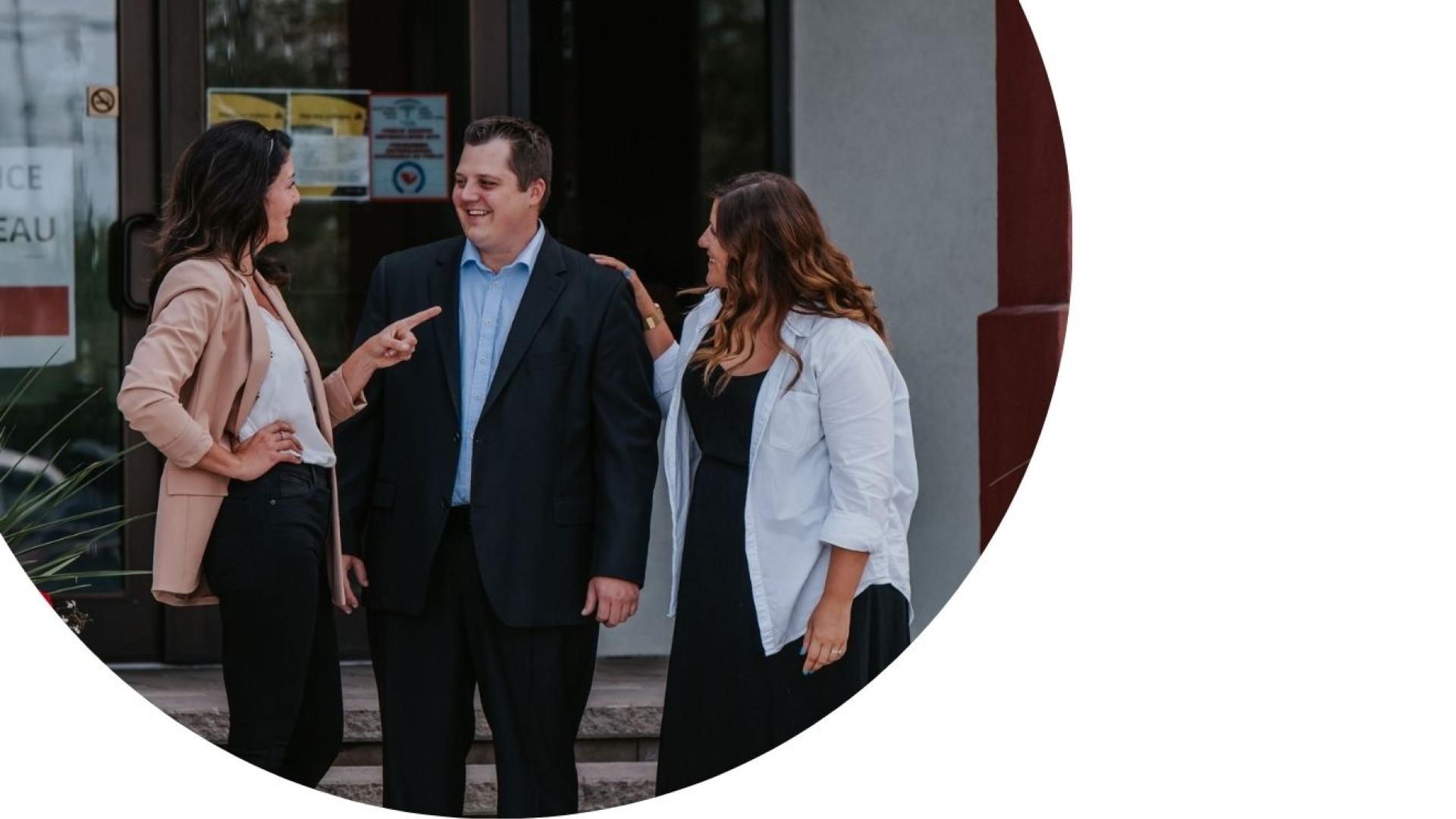 Three adults standing and talking outside a building entrance, engaged in conversation in a professional setting.