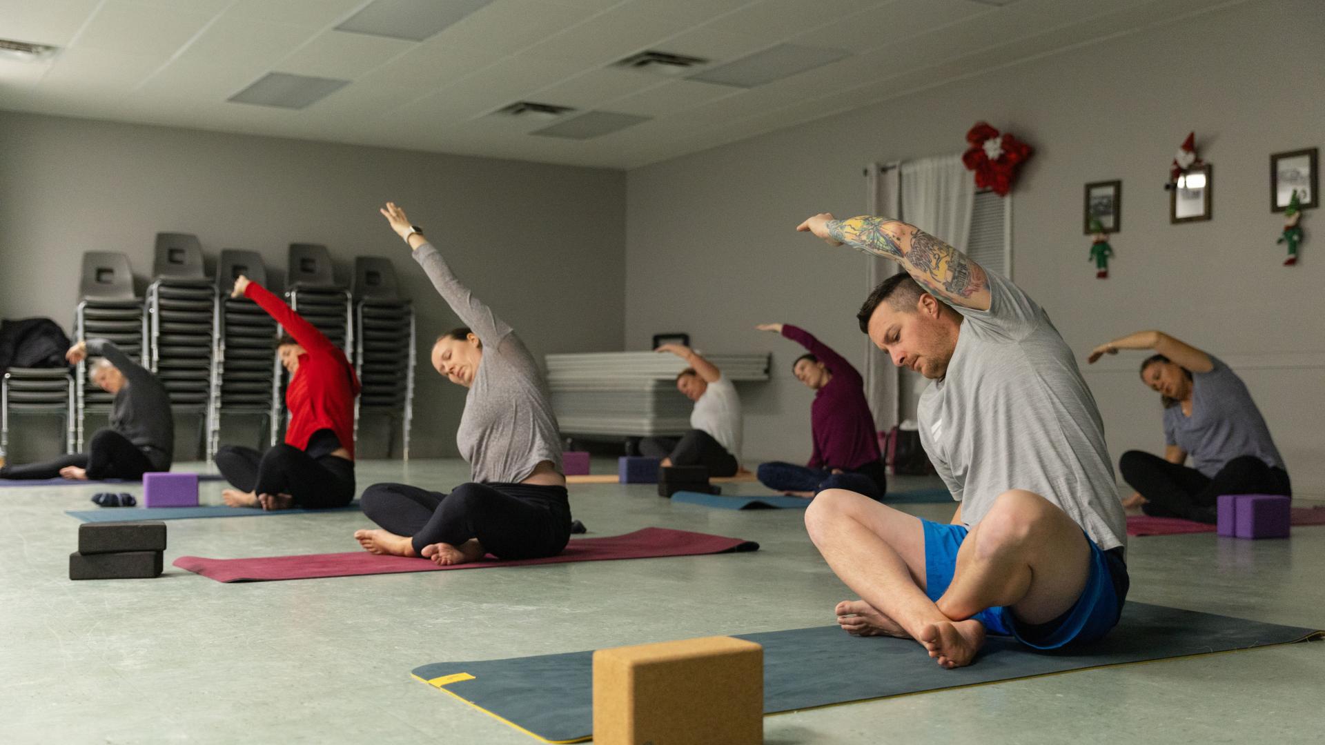 Adults participating in a fitness or yoga class, performing stretching exercises on mats in a community hall.
