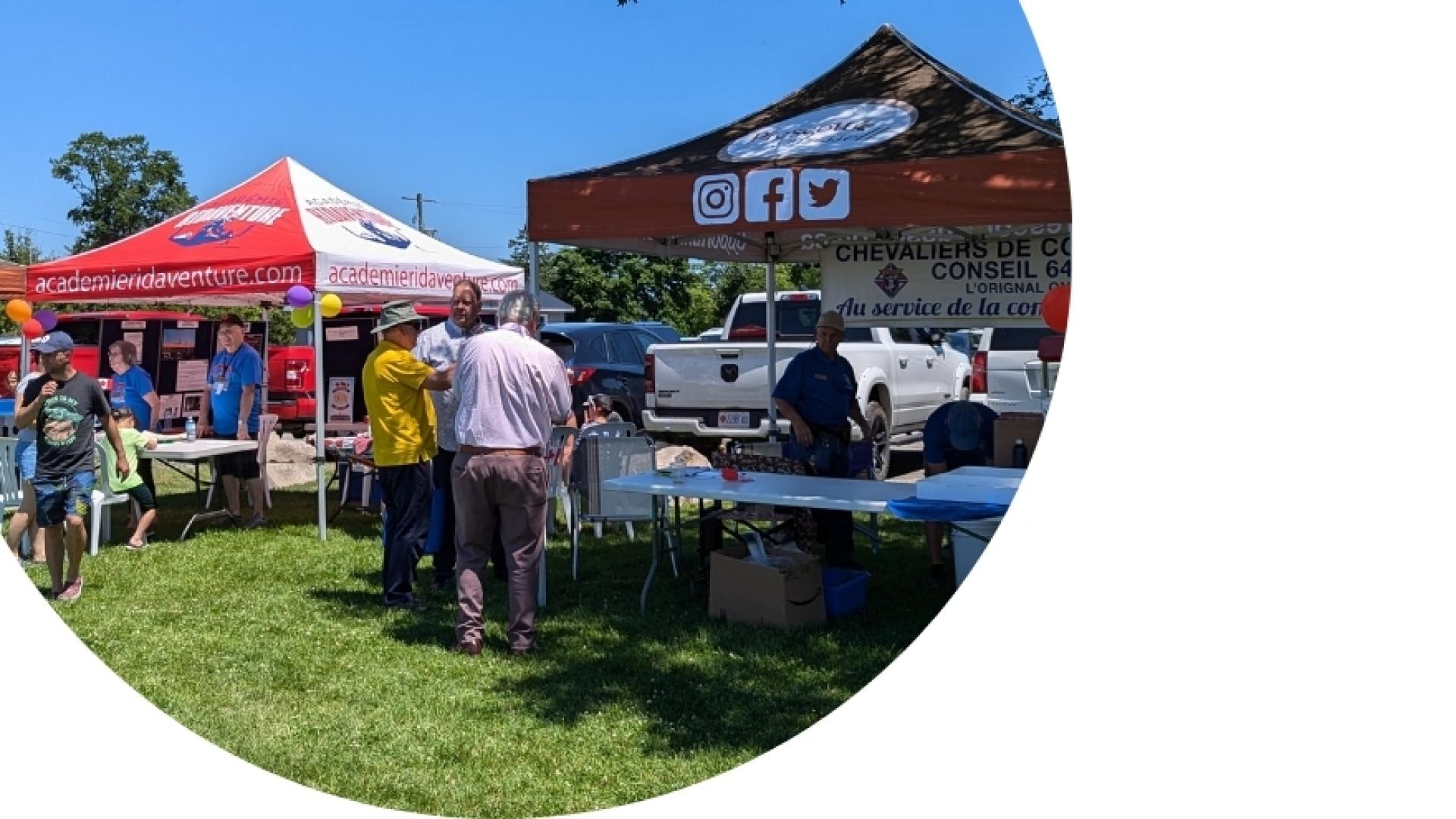 Community members visiting vendor booths and information tents at an outdoor community event held on a grassy field on a sunny day.