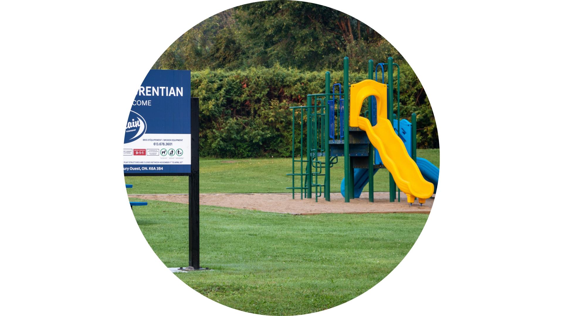 Playground featuring climbing structures and a slide, with open green space and a park sign in the foreground.