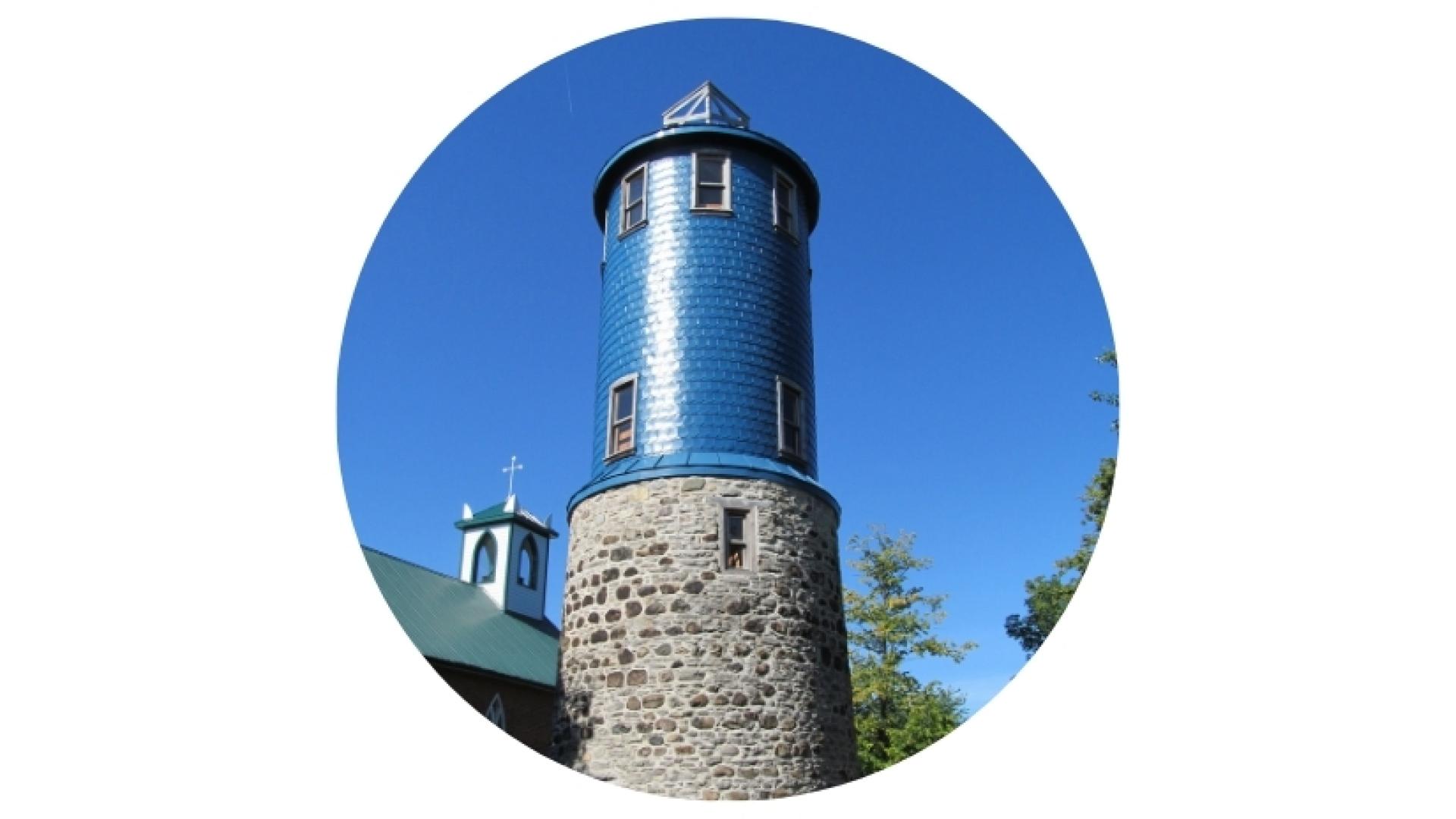 Historic stone tower with a blue cylindrical observation structure and conical roof, standing against a clear blue sky and surrounded by trees.