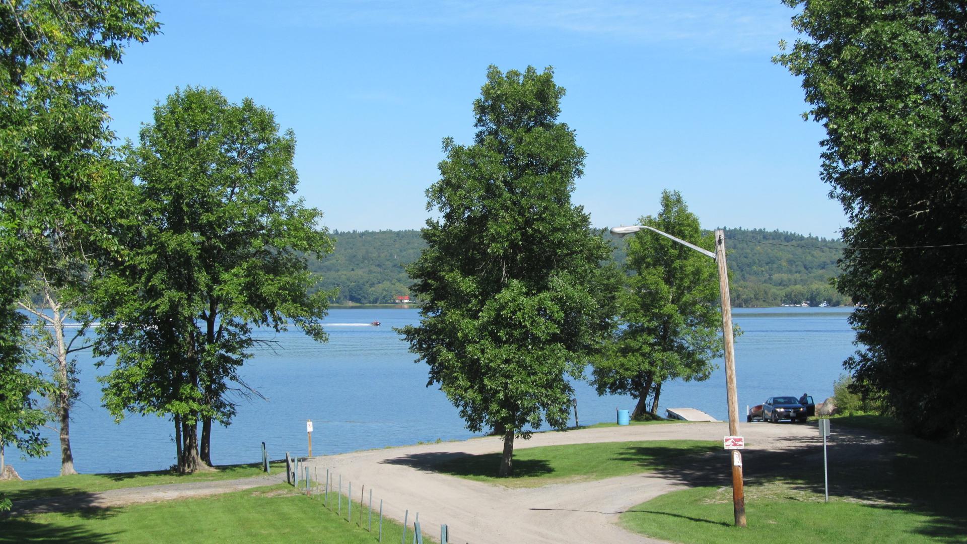 View of the Ottawa River from L'Orignal Park, featuring open green space, mature trees, a waterfront roadway, and boats on the water in the distance.