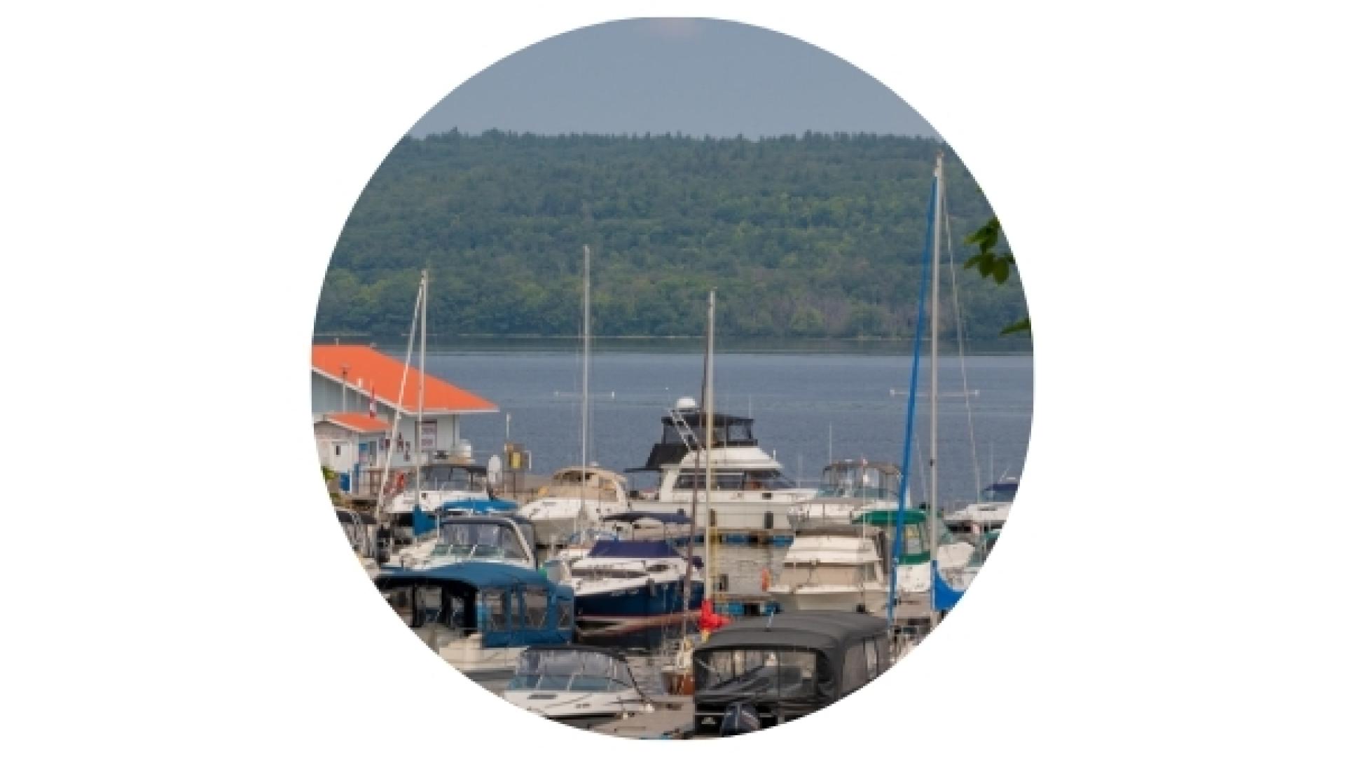 Boats docked at the L’Orignal Marina on the Ottawa River, with sailboats, recreational vessels, and a forested shoreline visible in the background.