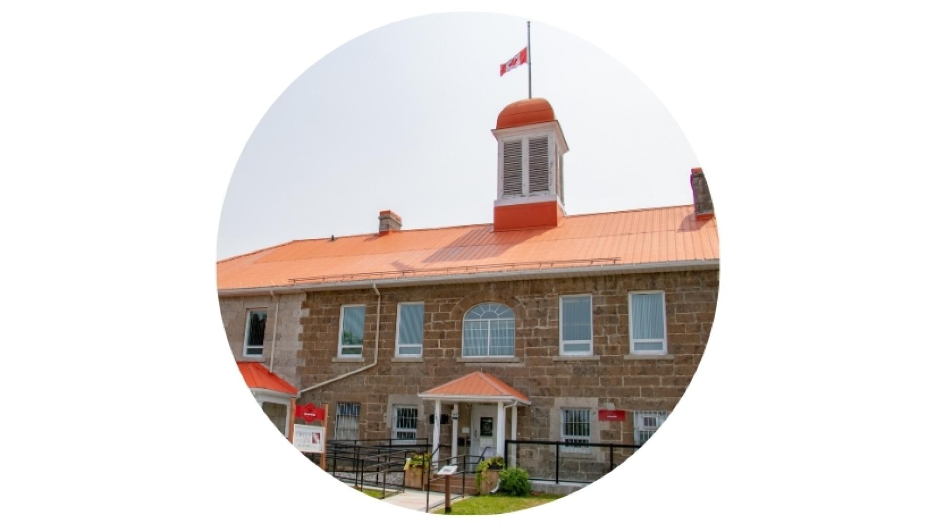 Historic stone building with a central cupola topped by a Canadian flag, featuring arched windows and a front entrance, shown in a circular image.