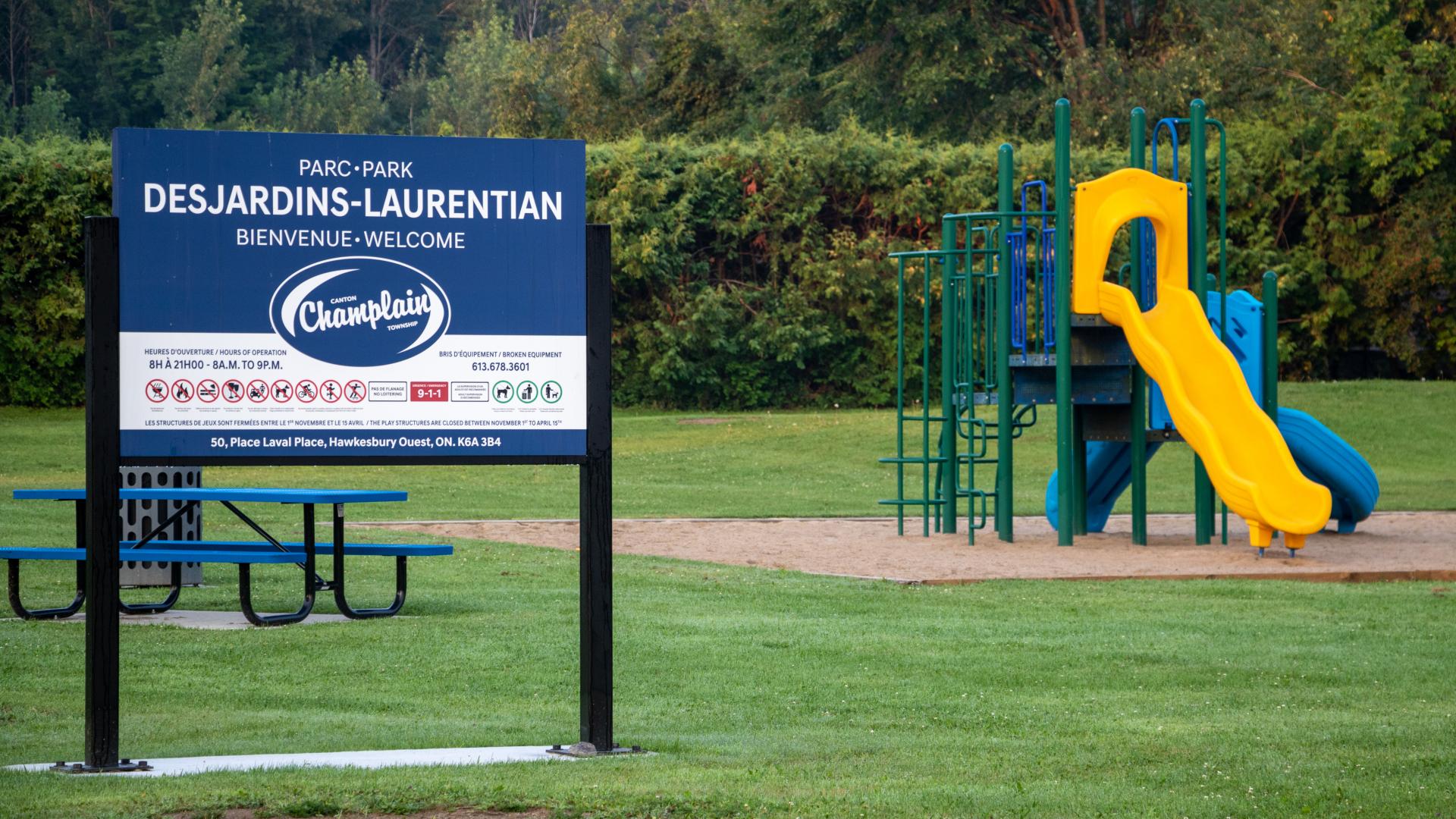 Playground featuring climbing structures and a slide, with open green space and a park sign in the foreground.