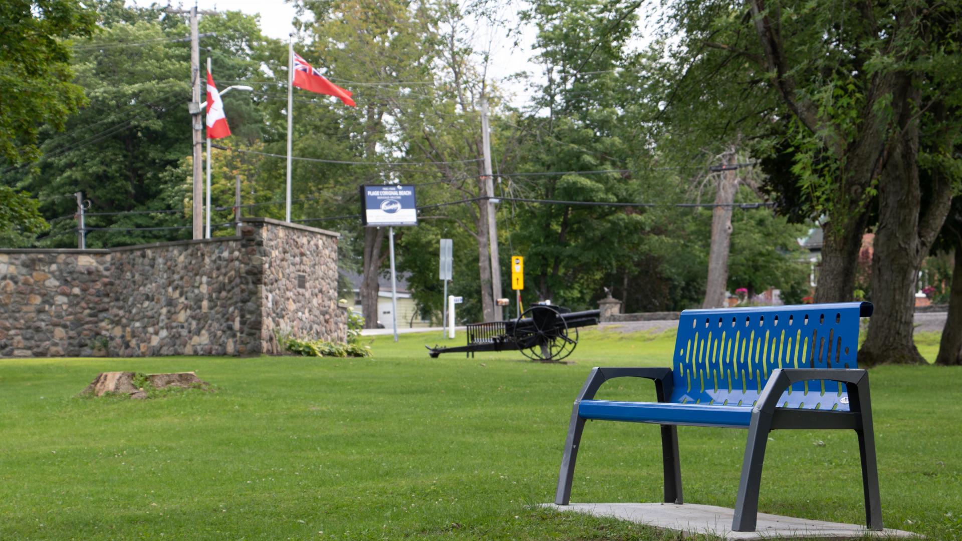 Bench and open green space in L'Orignal Park with trees and park amenities in the background.