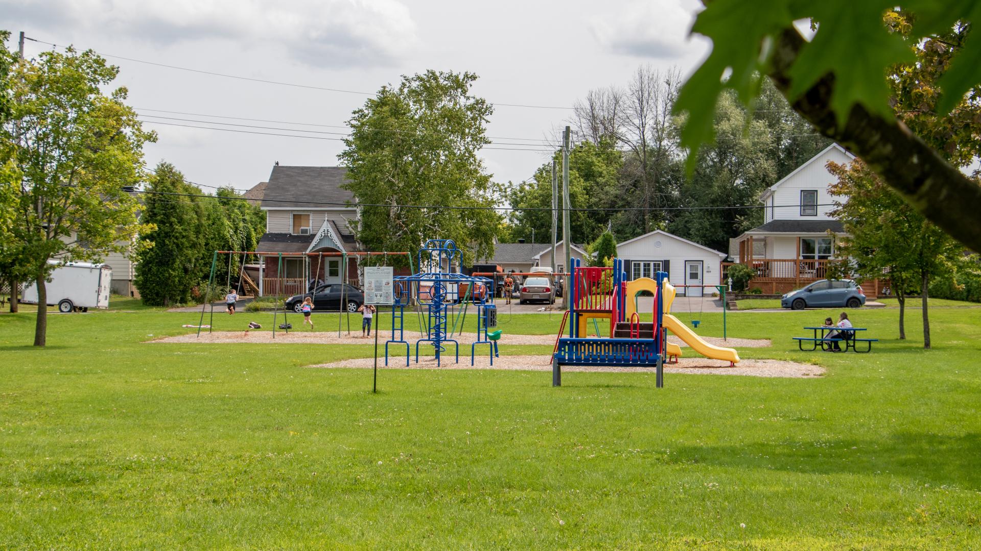 Neighbourhood park featuring open green space, a playground, and mature trees, with homes visible in the background.