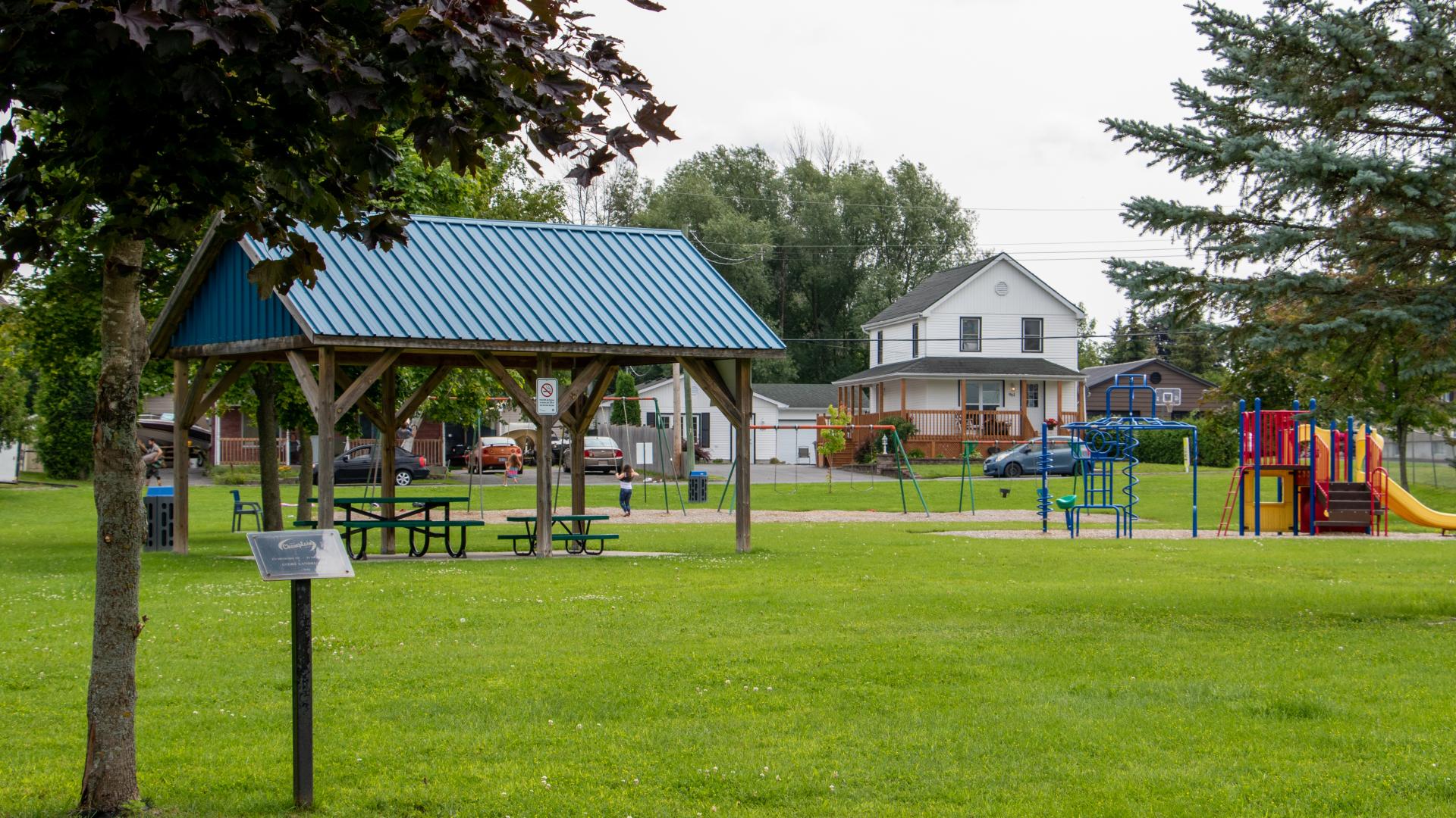 Community park featuring a covered picnic shelter, playground equipment, open green space, and mature trees.