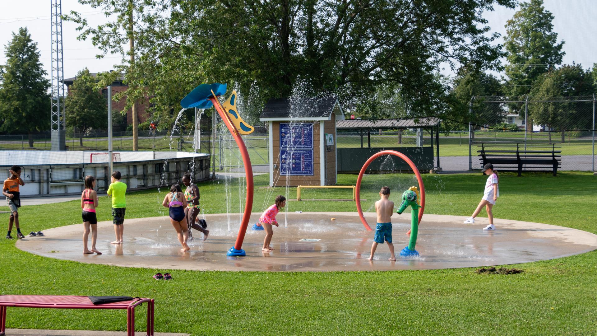 Children playing in a splash pad with interactive water features and spray arches in a community park.
