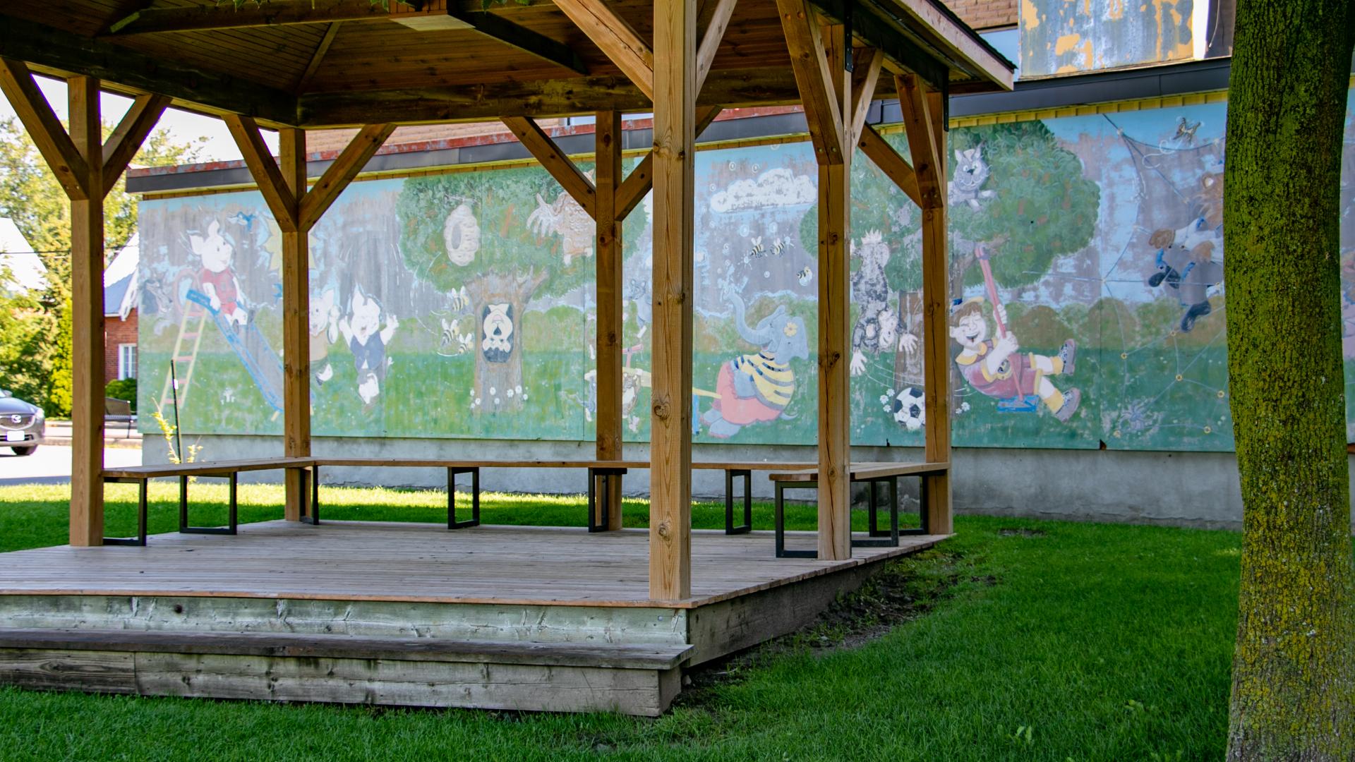 Wooden gazebo with benches in a park, set in front of a colourful community mural and surrounded by green space.