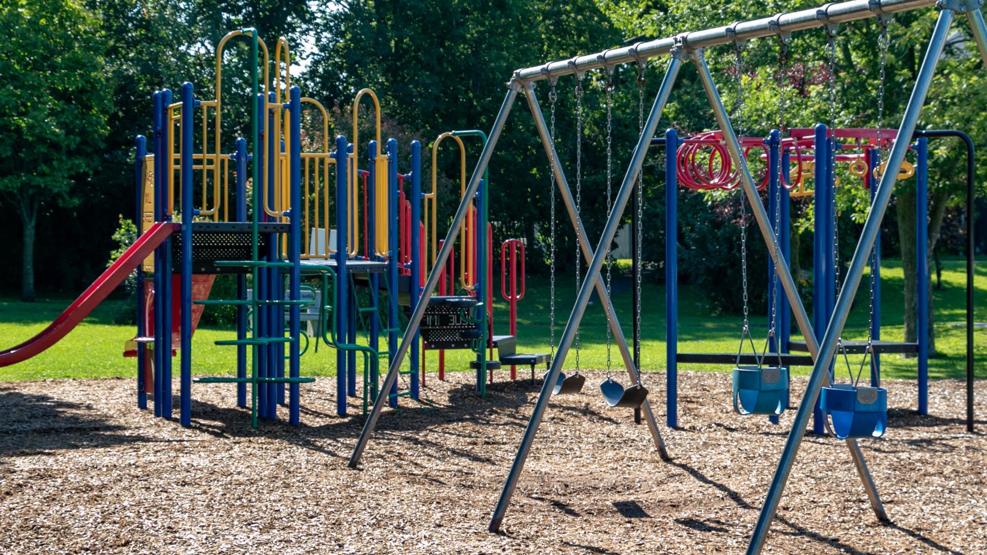 Playground with slides, climbing structures, and swings, surrounded by trees in a neighbourhood park.