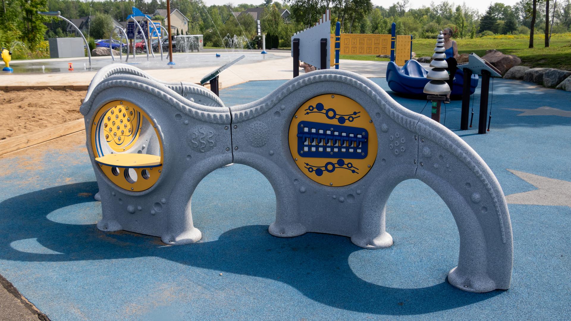 Accessible playground panel with musical, sensory and interactive play features at Sacha's Park, with the splash pad visible in the background.