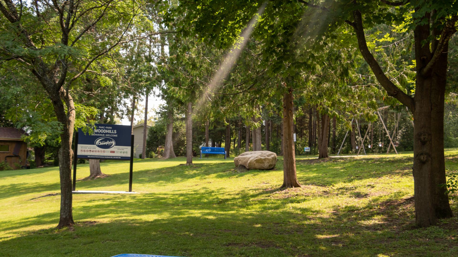 **Alt text:** Picnic table and open green space in Woodhills Park, with mature trees, park signage, and recreational facilities in the background.