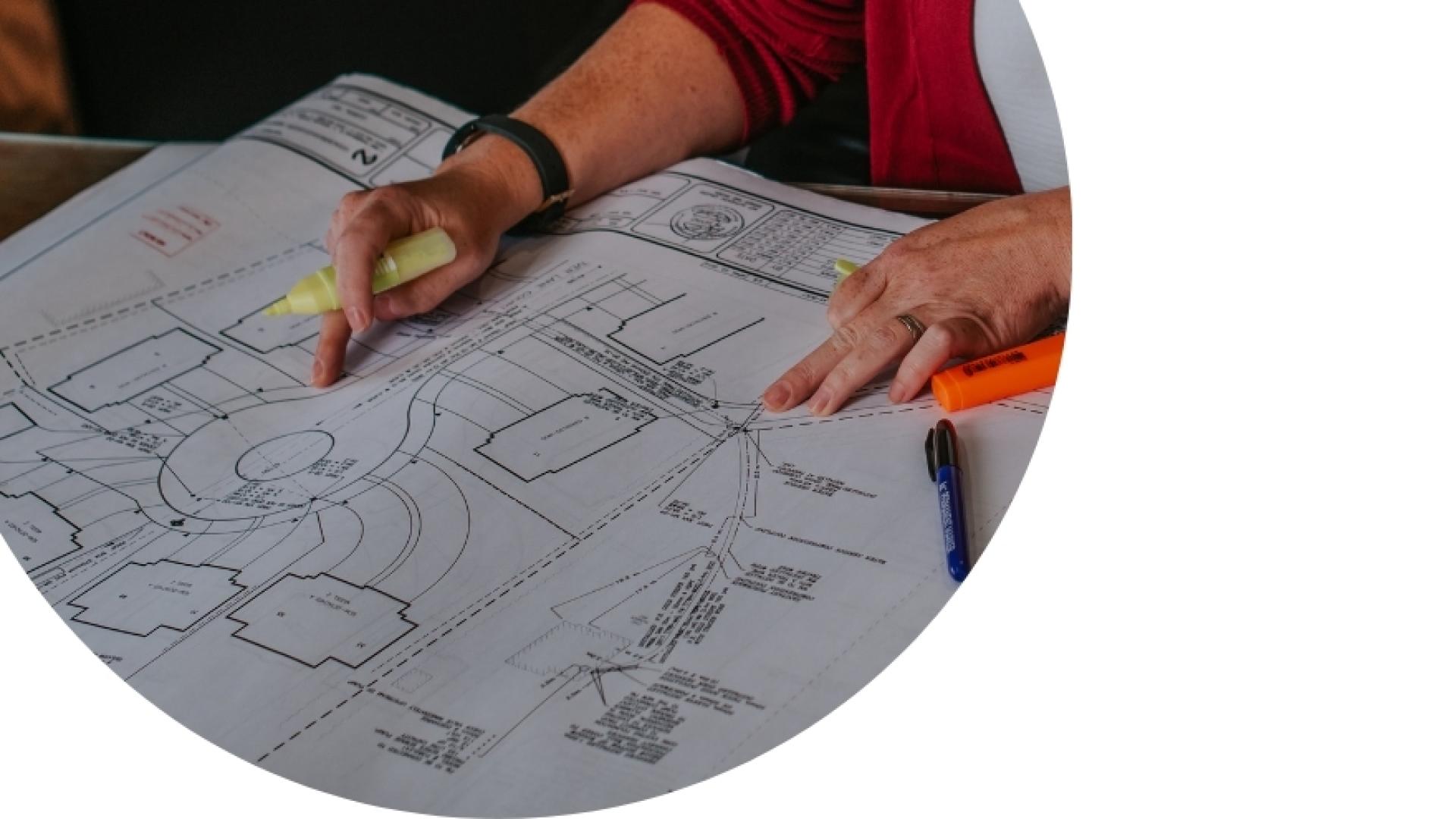 Person reviewing large architectural or site plans with a pencil and highlighter, marking details on a technical drawing spread across a table.