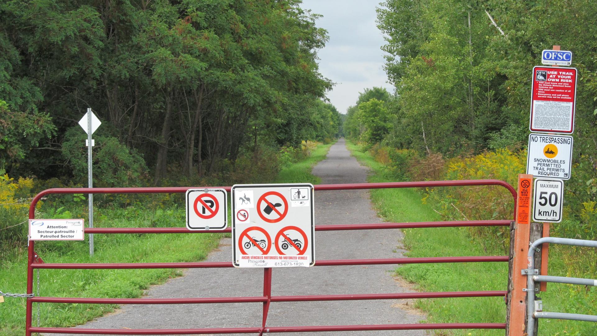 Entrance gate to the Prescott and Russell Recreational Trail, featuring trail signage and a paved pathway extending through a wooded landscape.