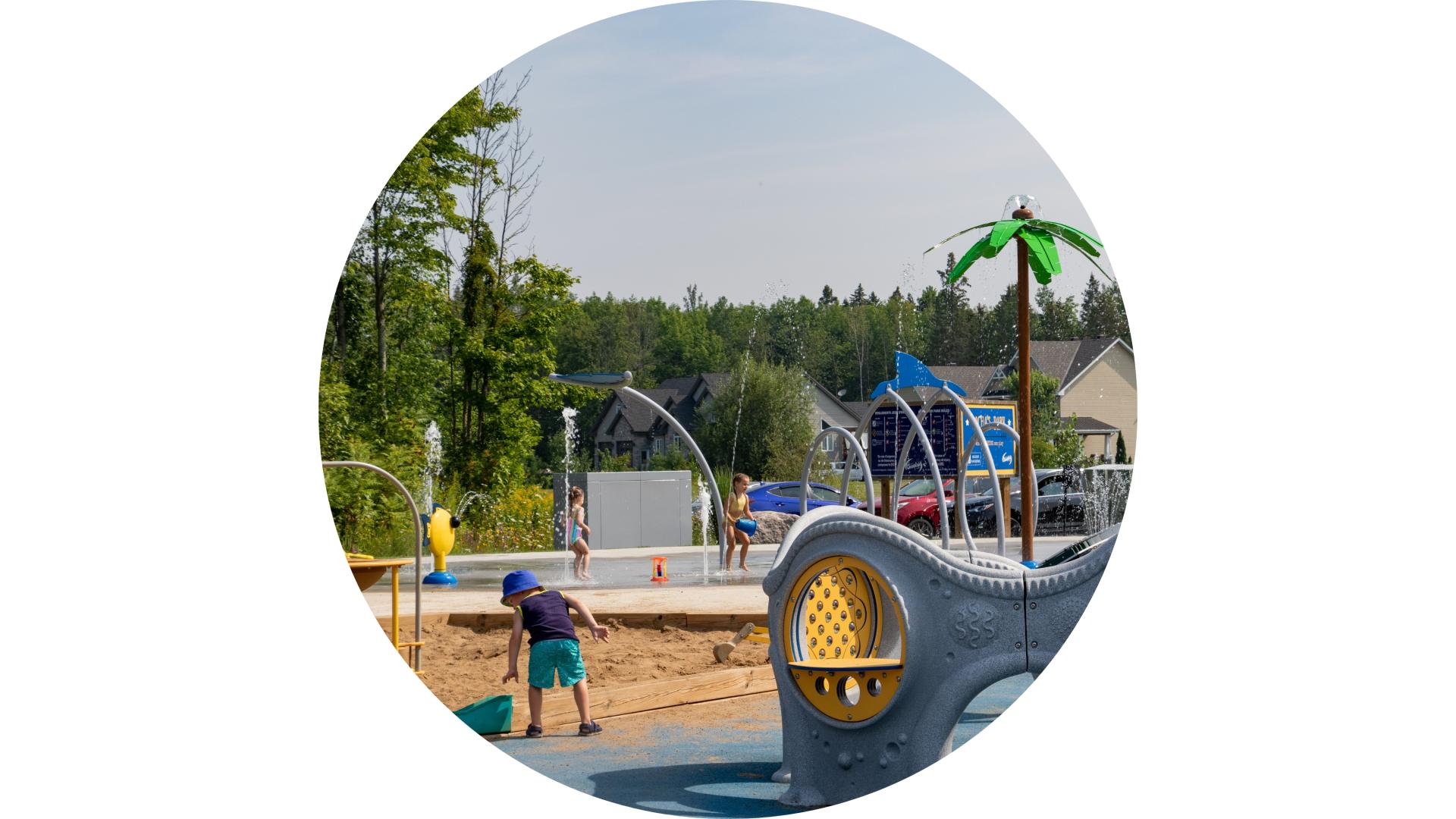 Children and adults enjoying a playground and splash pad area in a community park on a summer day.