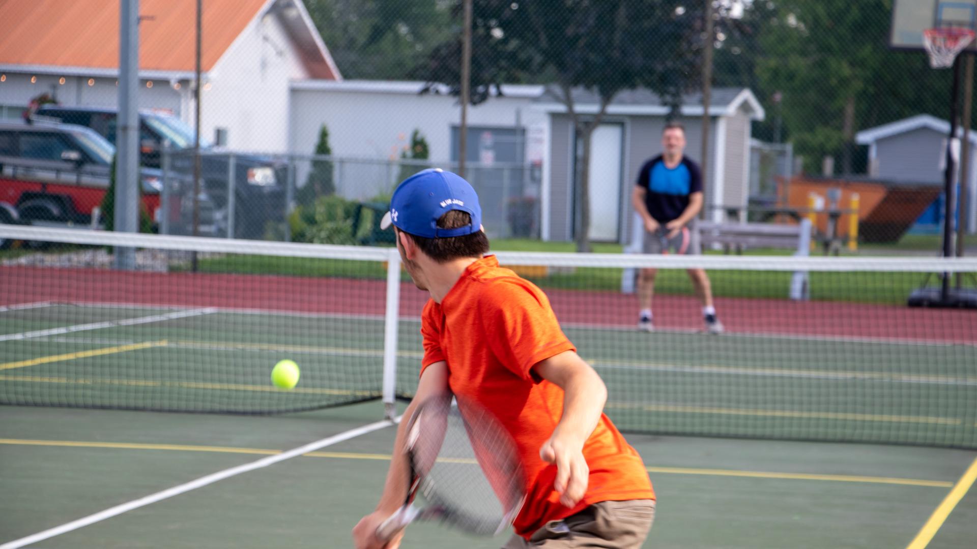 People playing tennis or pickleball on an outdoor court, with one player preparing to return the ball while others play in the background.