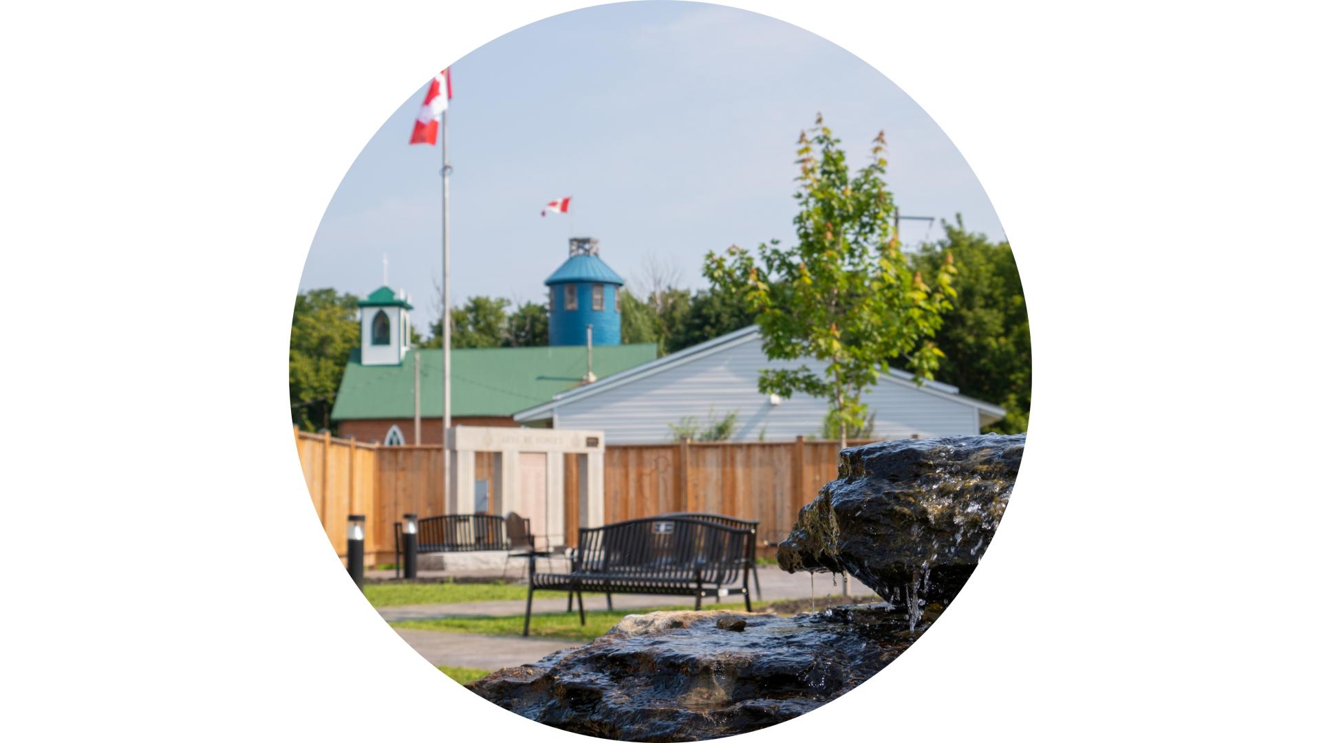 Town Square Park featuring benches, a decorative water fountain, Canadian flags, and a commemorative monument surrounded by landscaped green space.
