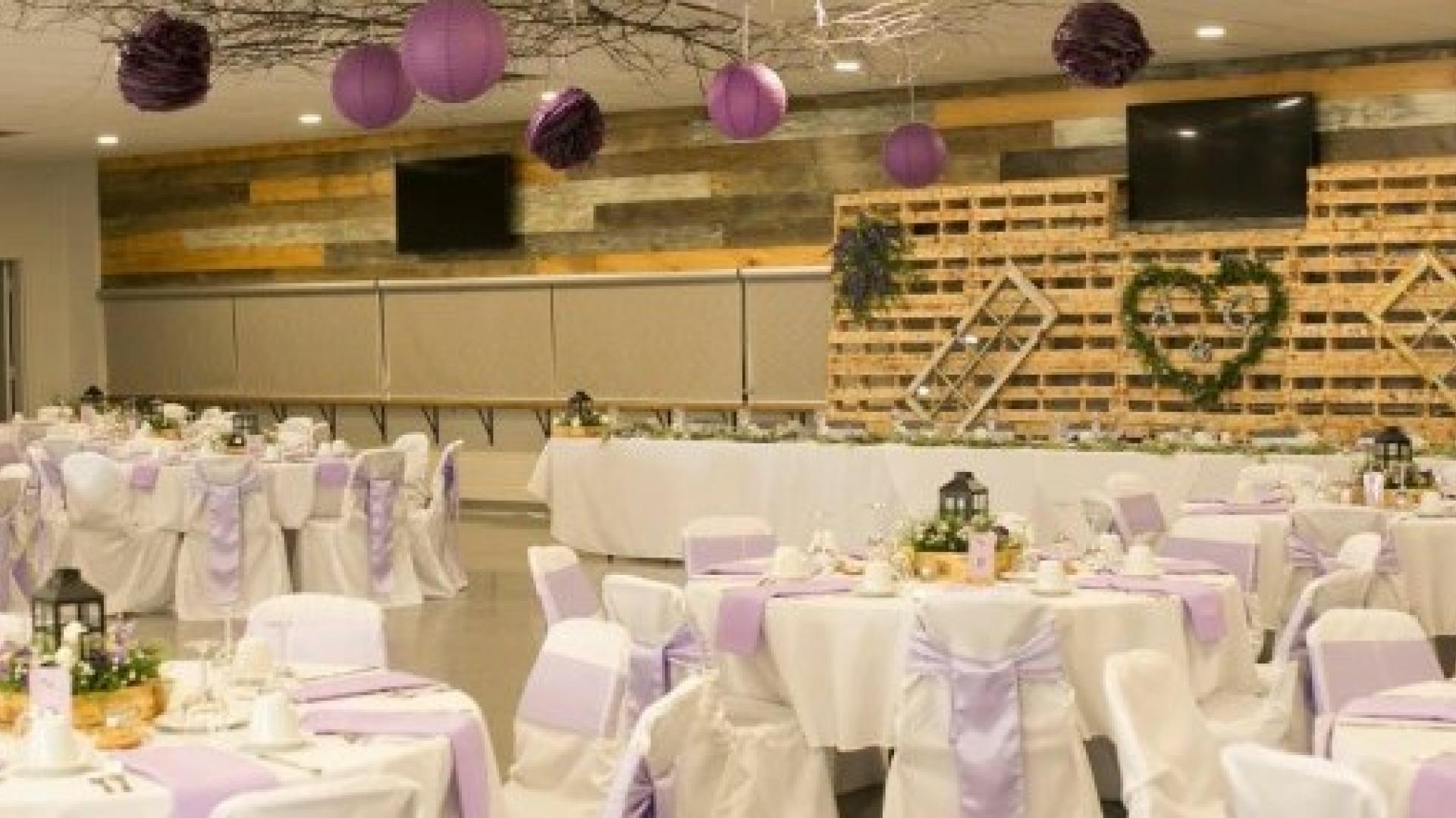 Community hall decorated for a wedding reception with round tables, white linens, floral centrepieces, and purple accents.