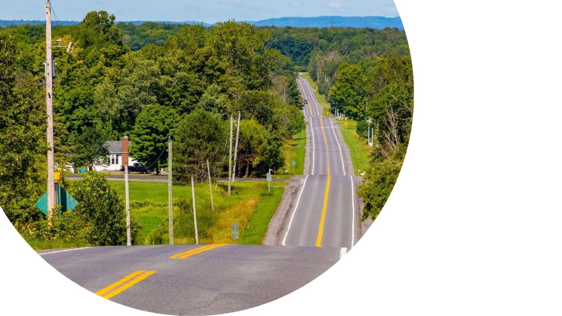 Rural two-lane road stretching through a forested landscape, with utility poles lining the roadside and rolling hills visible in the distance.