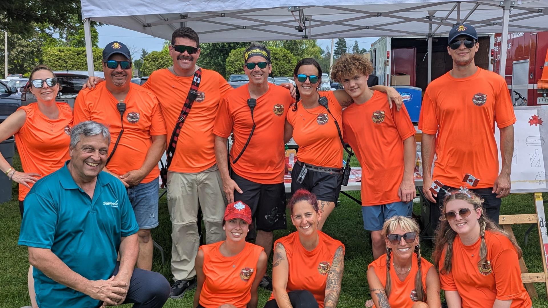 Group of volunteers wearing bright orange event T-shirts posing together on a grassy field during an outdoor community event. A white canopy tent and event booths are visible in the background.