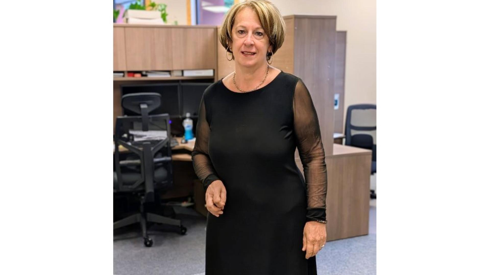 A woman wearing a black dress stands in an office, smiling at the camera.