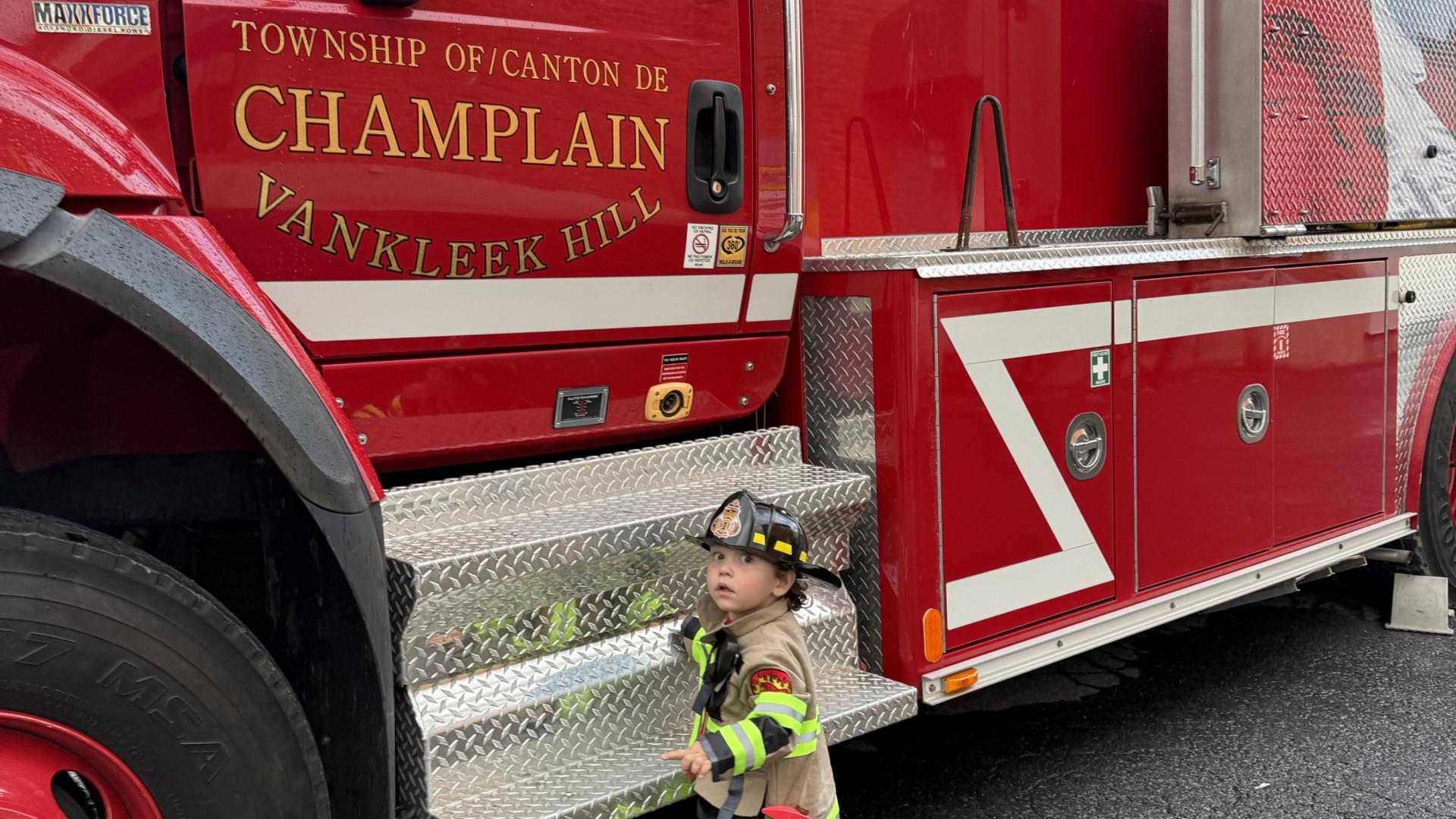 A child dressed as a firefighter standing in front of a red Vankleek Hill fire truck.