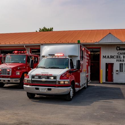 Red fire trucks and an ambulance parked outside a fire hall with an orange roof.