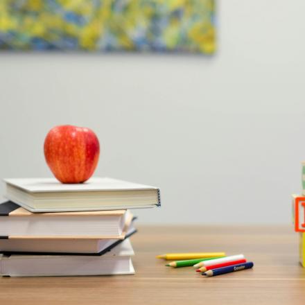 pile of books with an apple on top, with pens and cubes on a teacher's desk