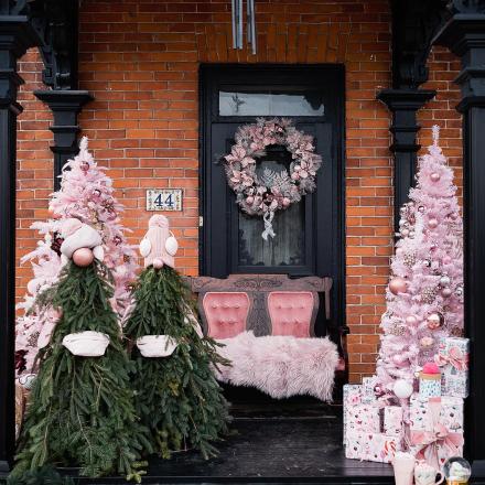 Pink holiday decorations on a porch with wreath and trees against a brick wall.