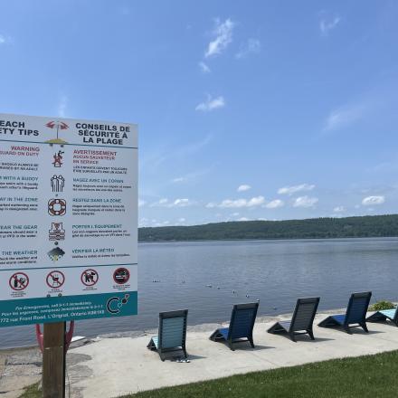 Beach safety rules sign beside the Ottawa River, with lounge chairs and the waterfront visible in the background.