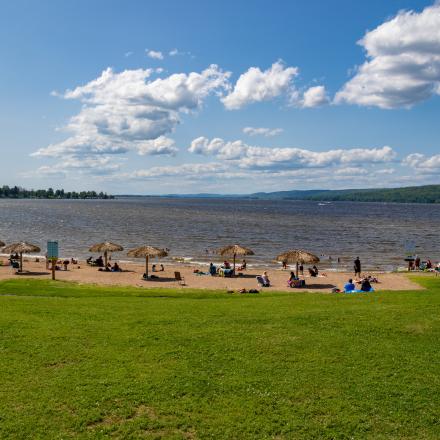 Wide view of L'Orignal Beach on a sunny day, showing a sandy shoreline, visitors relaxing and swimming, and the Ottawa River stretching into the distance under a blue sky with scattered clouds.