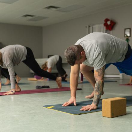 A group of adults participating in a fitness or yoga class, practicing stretching and balance exercises on mats in a community centre room.