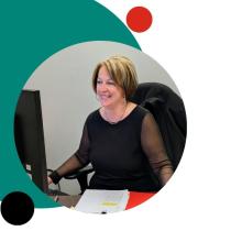 A woman sits at her desk in an office, smiling while working at a computer.